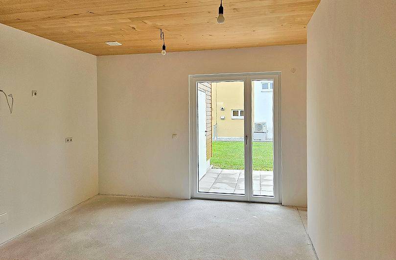 Geräumiger, unmöblierter Raum mit Holzbalkendecke und Schiebetür zur Terrasse und Rasenfläche.