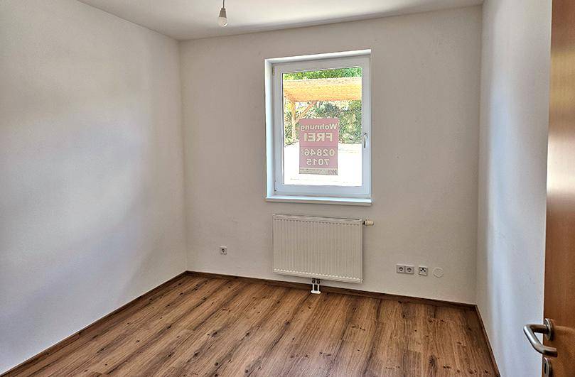 Helles Zimmer mit Laminatboden, Heizkörper und Fenster mit Blick auf den Carportbereich.