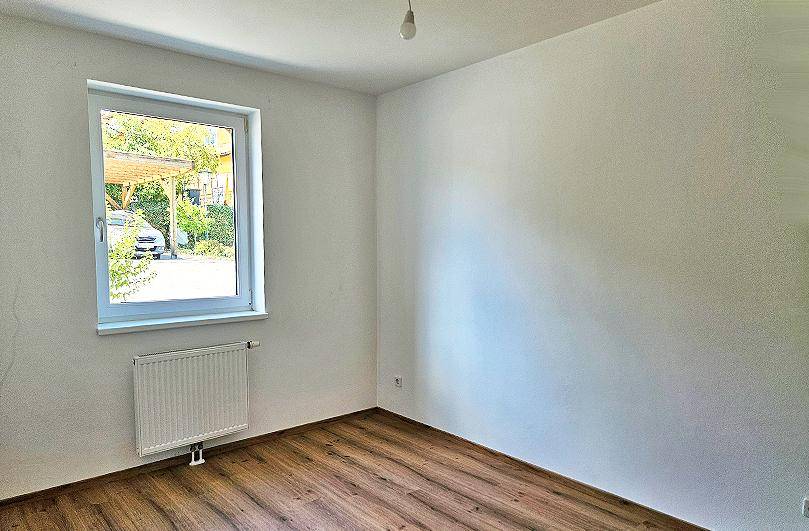 Leeres Zimmer mit Laminatboden, weiß gestrichenen Wänden und Fensterblick ins Grüne.