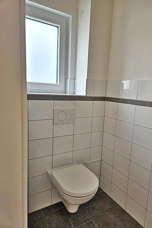 Modernes WC mit wandhängender Toilette, weißen Fliesen und einem kleinen Fenster.