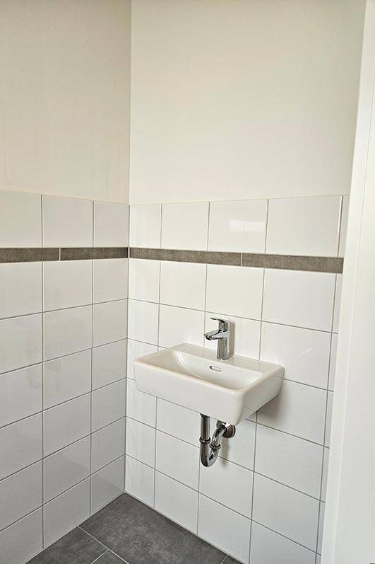 Kleines Waschbecken in einem gefliesten WC-Raum mit weißen Wandfliesen und dunklem Boden.