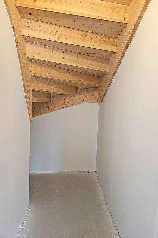 Unfertiger Bereich unter einer Holztreppe mit weißen Wänden und Betonboden.