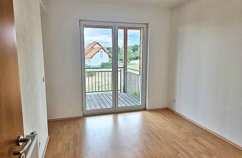 Leeres Zimmer mit Laminatboden und Zugang zu einem kleinen Balkon mit Blick ins Grüne.