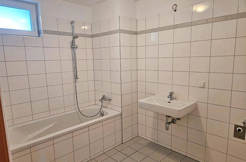 Badezimmer mit Badewanne, Waschbecken und weißen Fliesen, inklusive Fenster für Tageslicht.