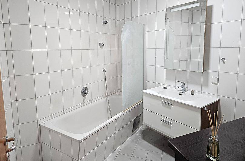 Badezimmer mit weißen Fliesen, Badewanne mit Duschwand und modernem Waschtisch mit Spiegelschrank.