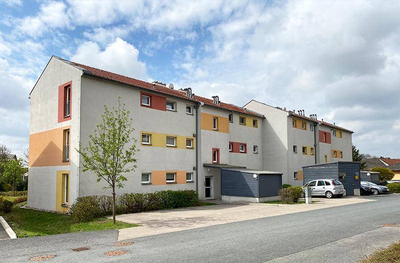 Mehrfamilienhaus mit moderner Architektur, farbiger Fassadengestaltung und Satteldach, umgeben von Grünflächen und Parkplätzen.