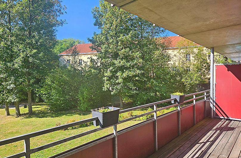 Geräumiger Balkon mit Holzboden und Blick ins Grüne sowie auf ein Gebäude.