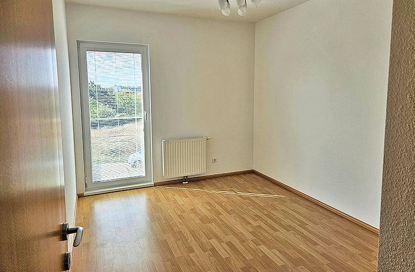 Helles Zimmer mit Parkettboden, Heizkörper und Fenstertür mit Jalousien.