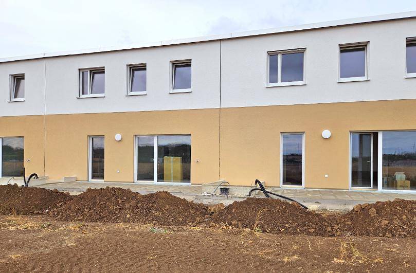Fassade eines Neubaus mit großen Fenstern und Terrassentüren, umgeben von Erdhügeln.