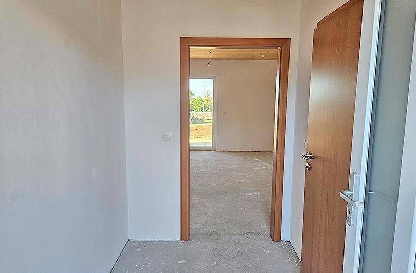 Blick von einem Flur in einen hellen Raum mit Zugang zum Außenbereich und unfertigem Boden.