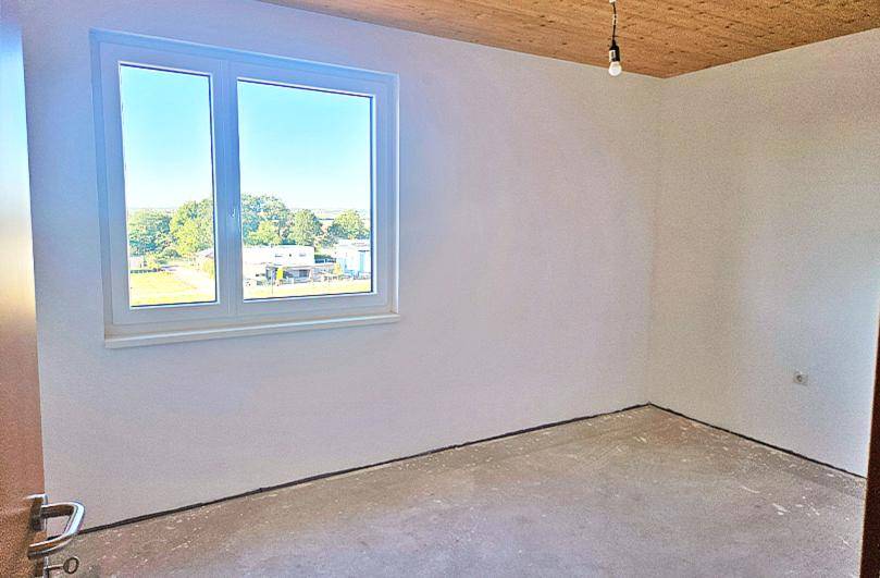 Unfertiges Zimmer mit großem Fenster, Holzbalkendecke und Blick ins Grüne.