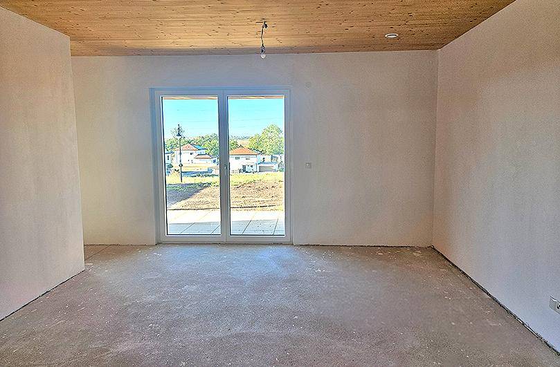 Heller, unfertiger Wohnraum mit Holzbalkendecke, großem Fenster und Zugang zum Gartenbereich.