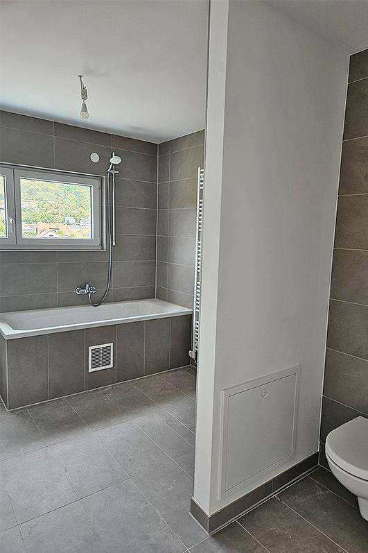 Modernes Badezimmer mit grauen Fliesen, Badewanne und Fenster für natürliches Licht.