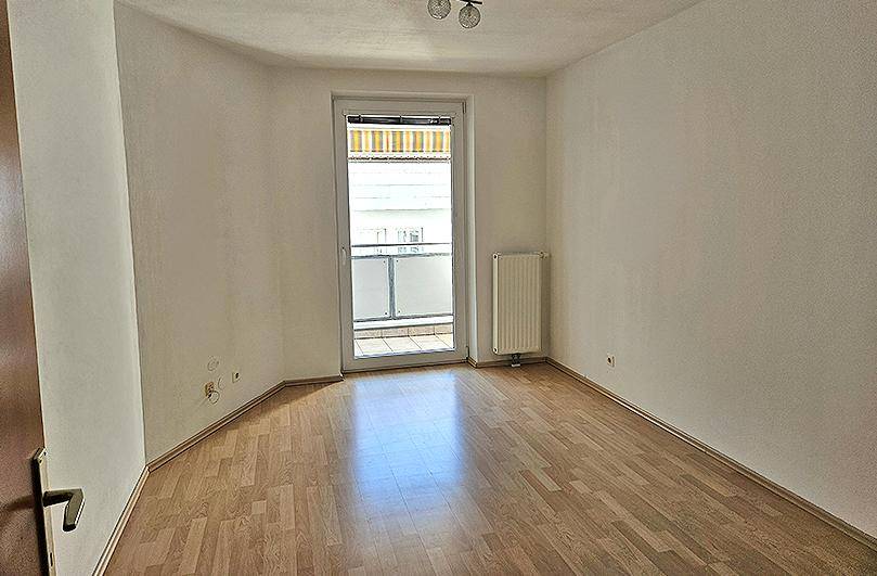 Leeres Schlafzimmer mit Laminatboden, weißen Wänden und direktem Zugang zu einem kleinen Balkon.
