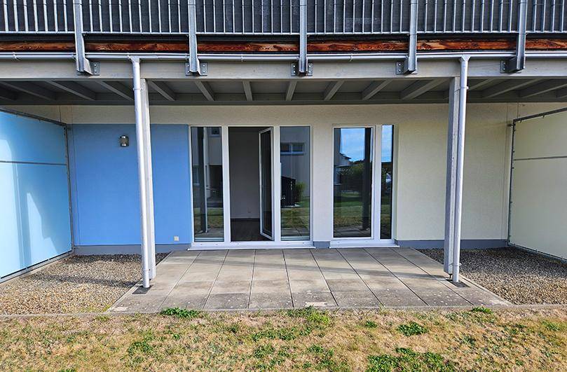 Erdgeschoss-Terrasse mit Betonplatten und blauem Sichtschutz unter einem darüberliegenden Balkon.