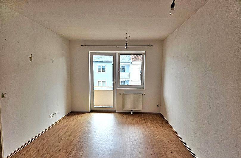Geräumiges Wohnzimmer mit Laminatboden, weißen Wänden und einem großen Fenster mit Balkonzugang.