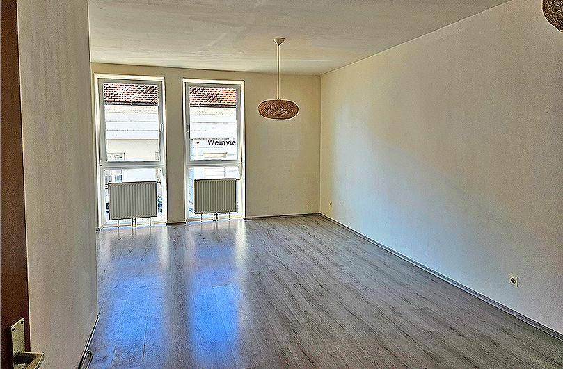 Geräumiges Wohnzimmer mit Laminatboden, zwei großen Fenstern und einem Deckenlicht.