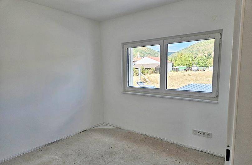 Leerer Raum mit weißen Wänden, unfertigem Boden und Fenster mit Blick ins Grüne.