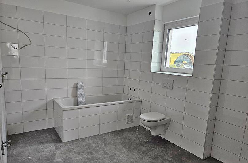 Unfertiges Badezimmer mit weißen Fliesen, eingebauter Badewanne und wandhängendem WC.