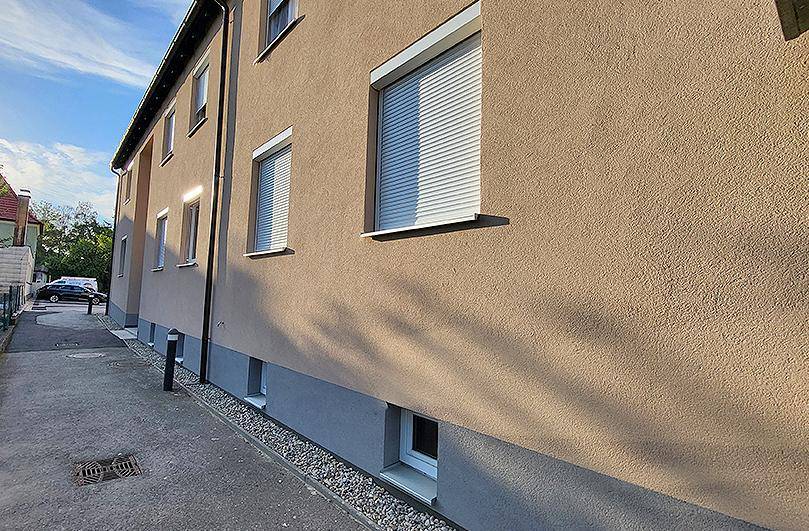 Seitenansicht des Gebäudes mit beiger Fassade, Fenstern und geschlossenen Rollläden.