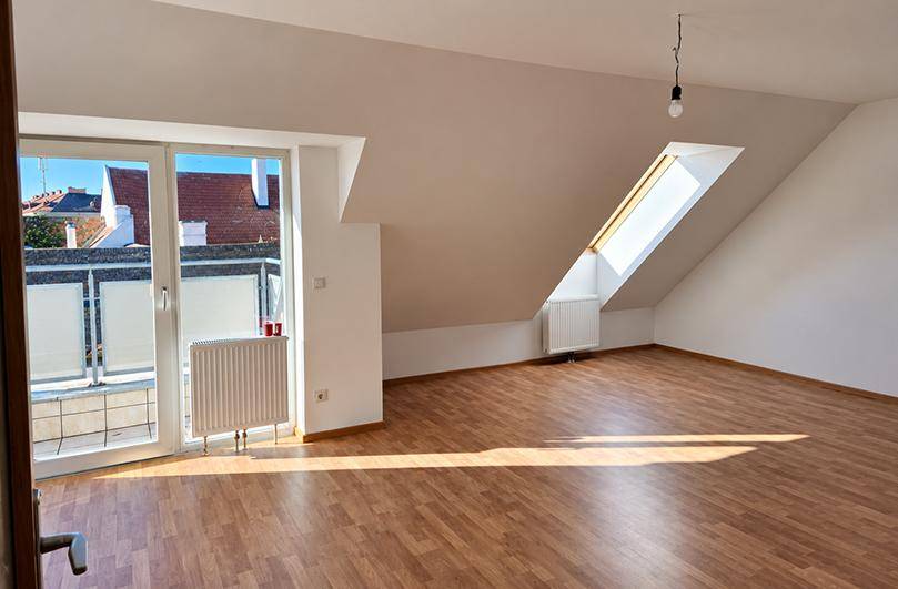 Lichtdurchfluteter Dachgeschossraum mit Holzboden, Dachfenster und Zugang zu einem sonnigen Balkon.