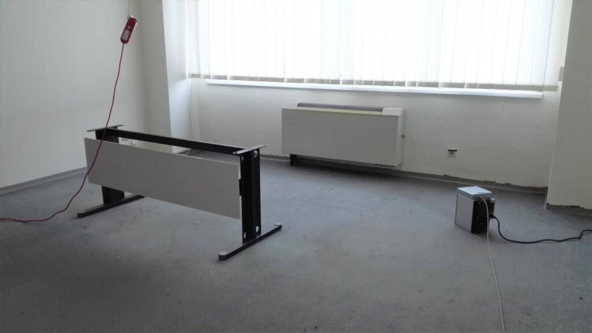 Leeres Büro mit grauem Boden, weißen Wänden, großen Fenstern und einem Heizkörper, mit Kabeln am Boden.