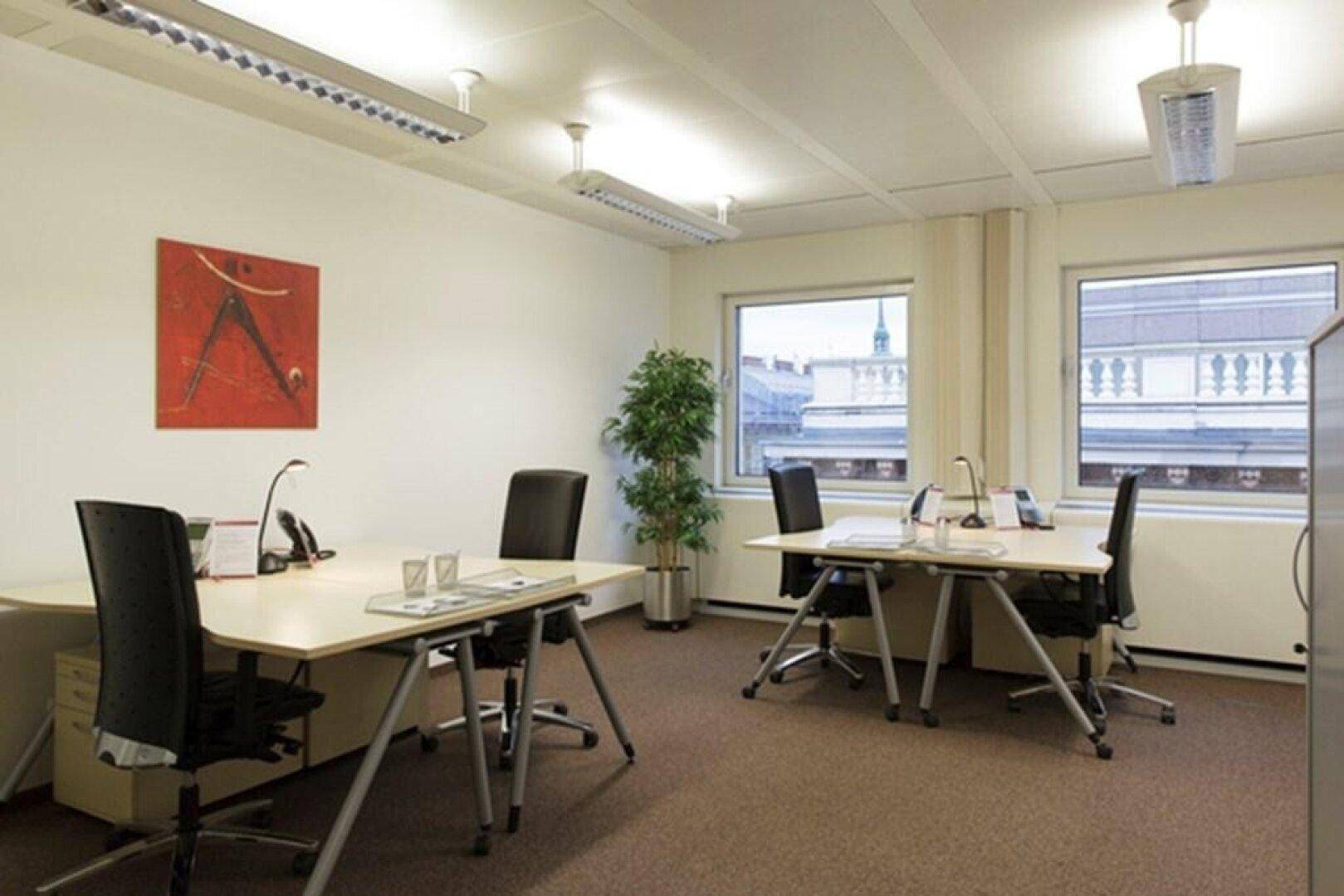 Helles Büro mit zwei Arbeitsplätzen, ergonomischen Stühlen und Blick auf die umliegenden Gebäude durch die Fenster.