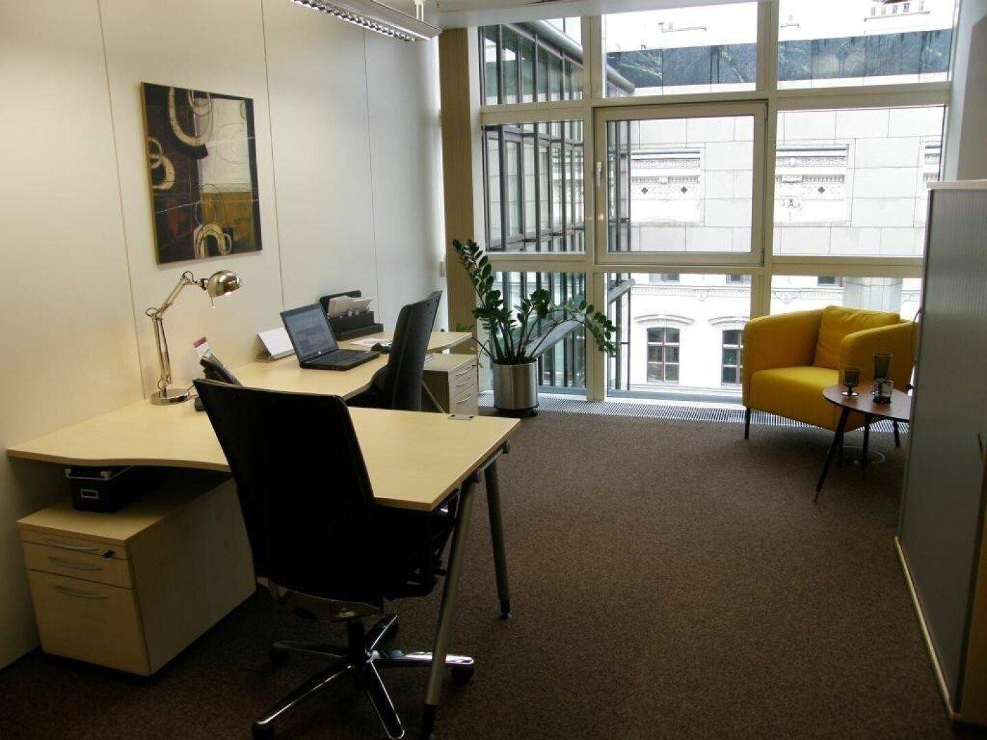 Modernes Büro mit großem Schreibtisch, gelbem Sessel und raumhohen Fenstern mit Blick auf die Nachbargebäude.