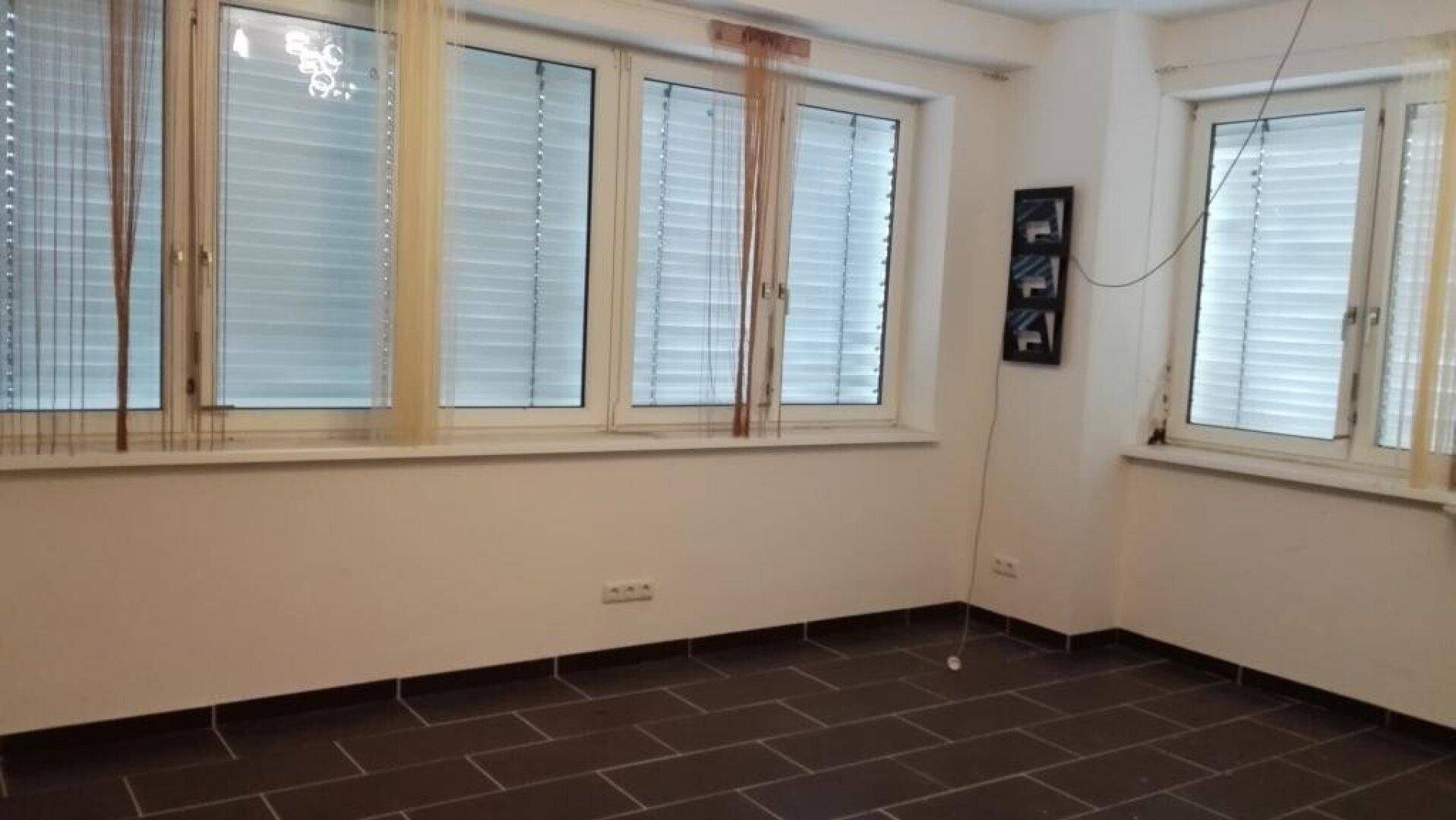 Leerer Raum mit dunklen Fliesen, weißen Wänden und mehreren Fenstern mit Jalousien.