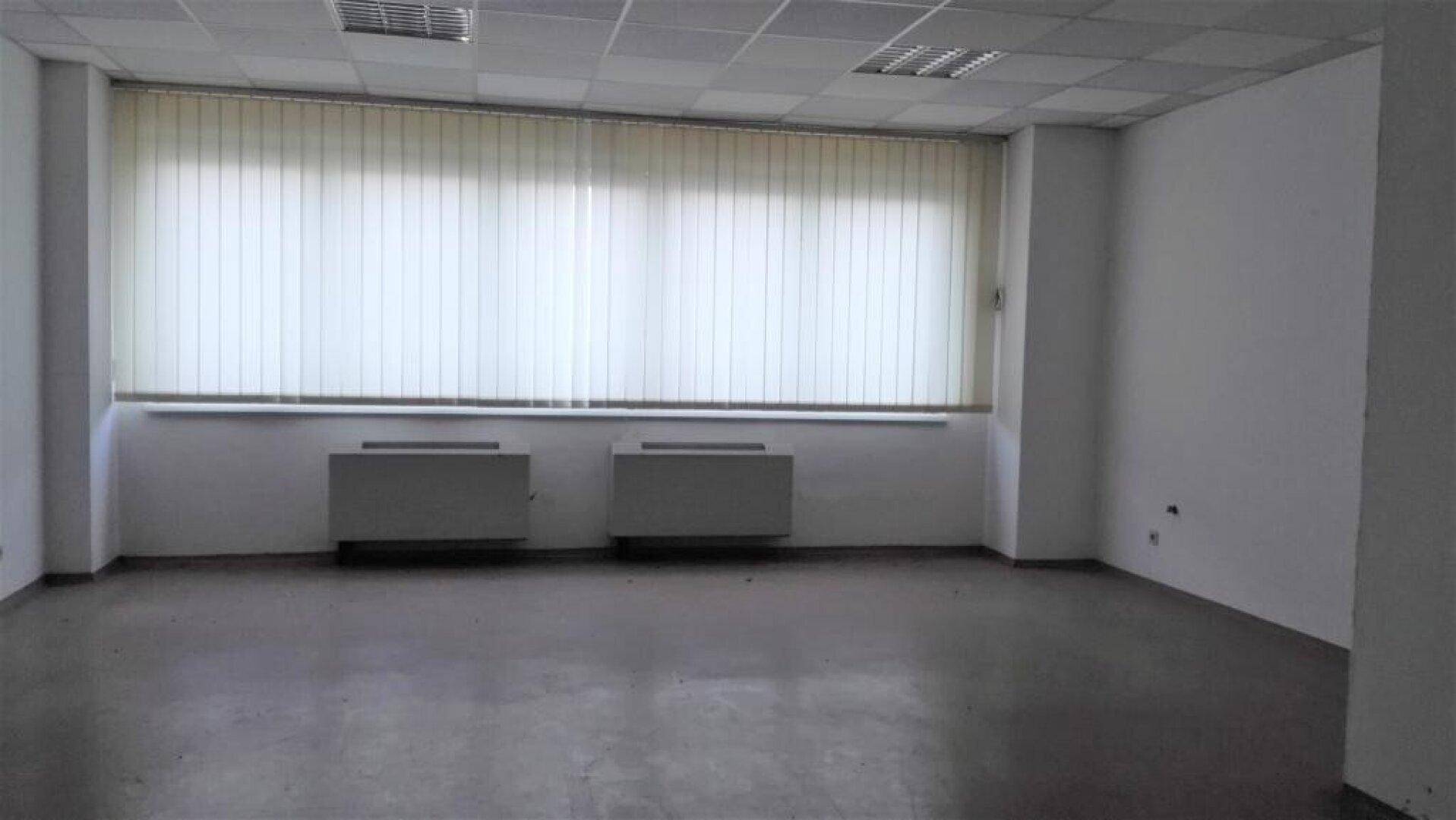 Leeres Büro mit grauem Boden, weißen Wänden, großen Fenstern mit Jalousien und zwei Heizkörpern.