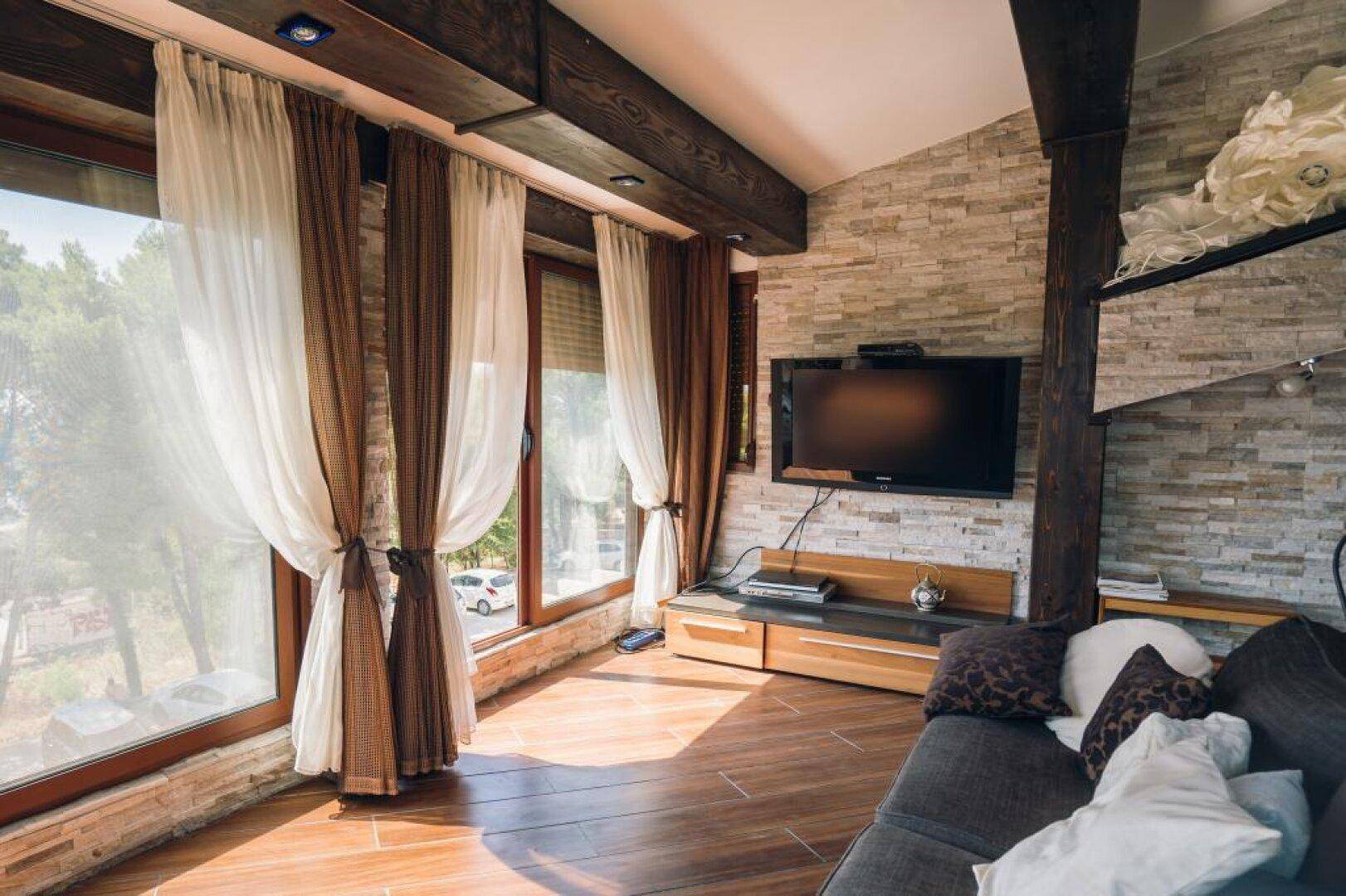 Helles Wohnzimmer mit großen Fenstern, Holzbalken und einem Fernseher an der Steinwand.