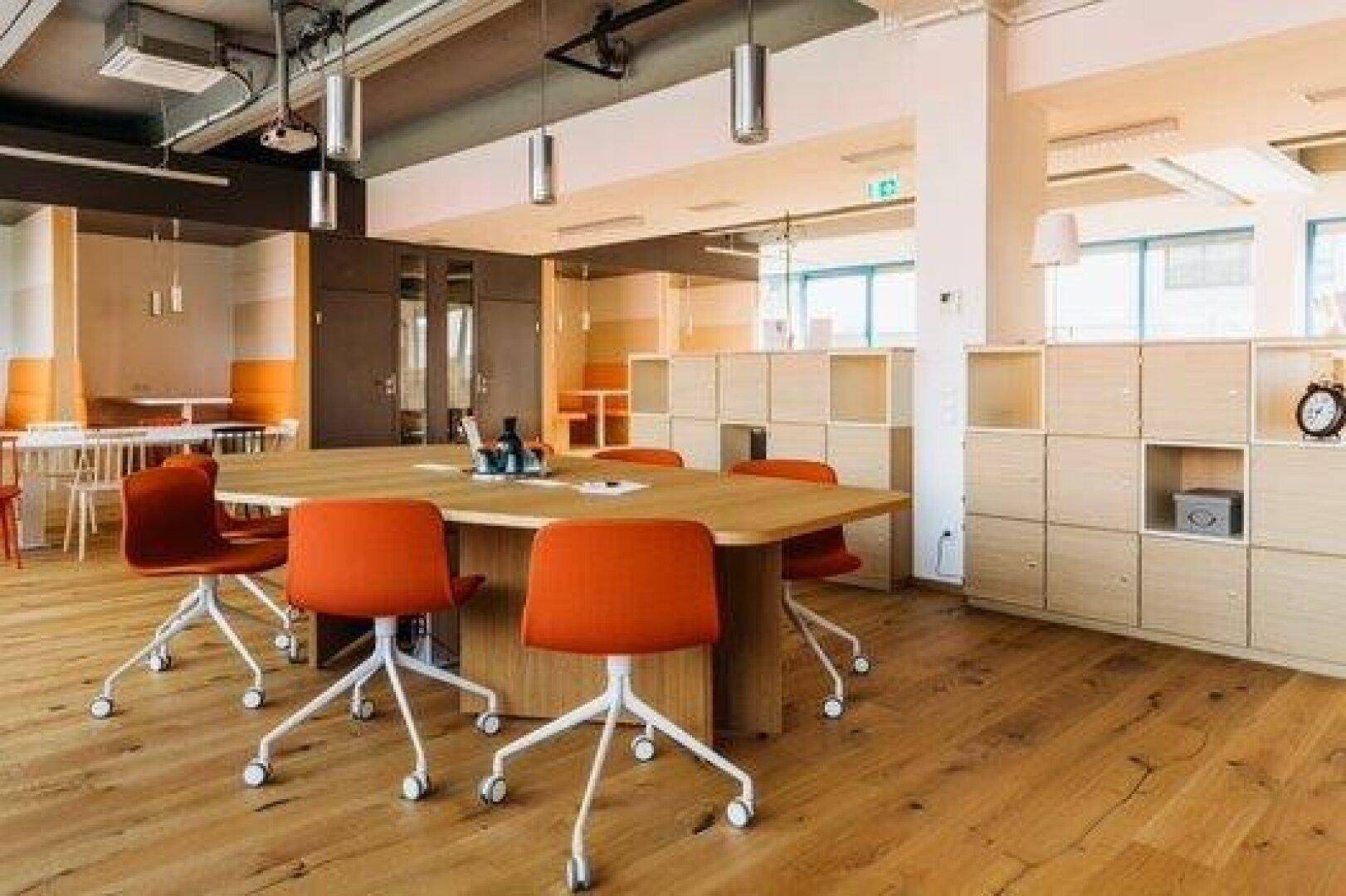 Großer Konferenzraum mit Holztisch, orangefarbenen Stühlen und Blick auf den offenen Bürobereich.