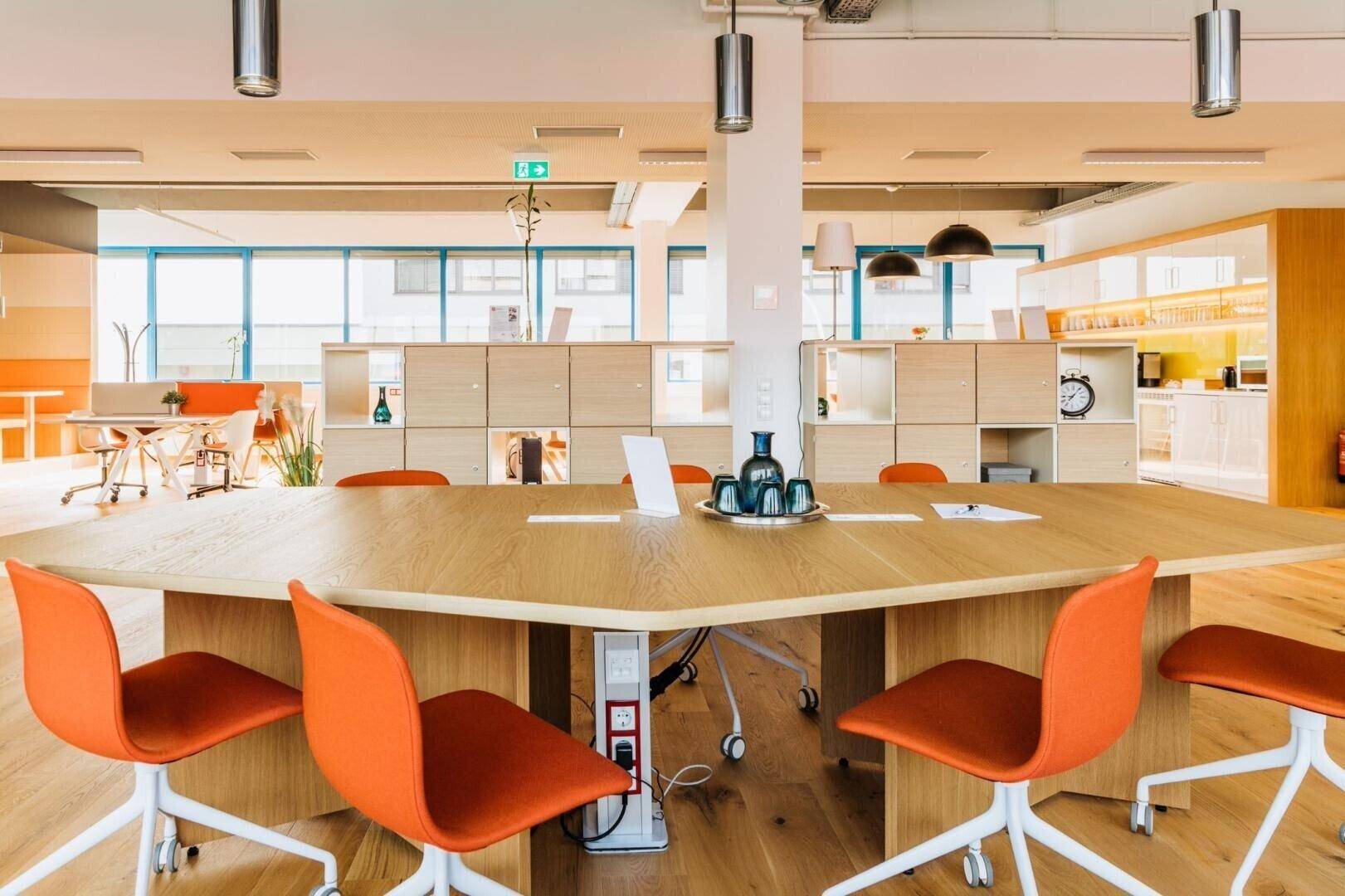 Moderner Konferenzraum mit großem Holztisch, orangefarbenen Stühlen und Blick auf den offenen Bürobereich.