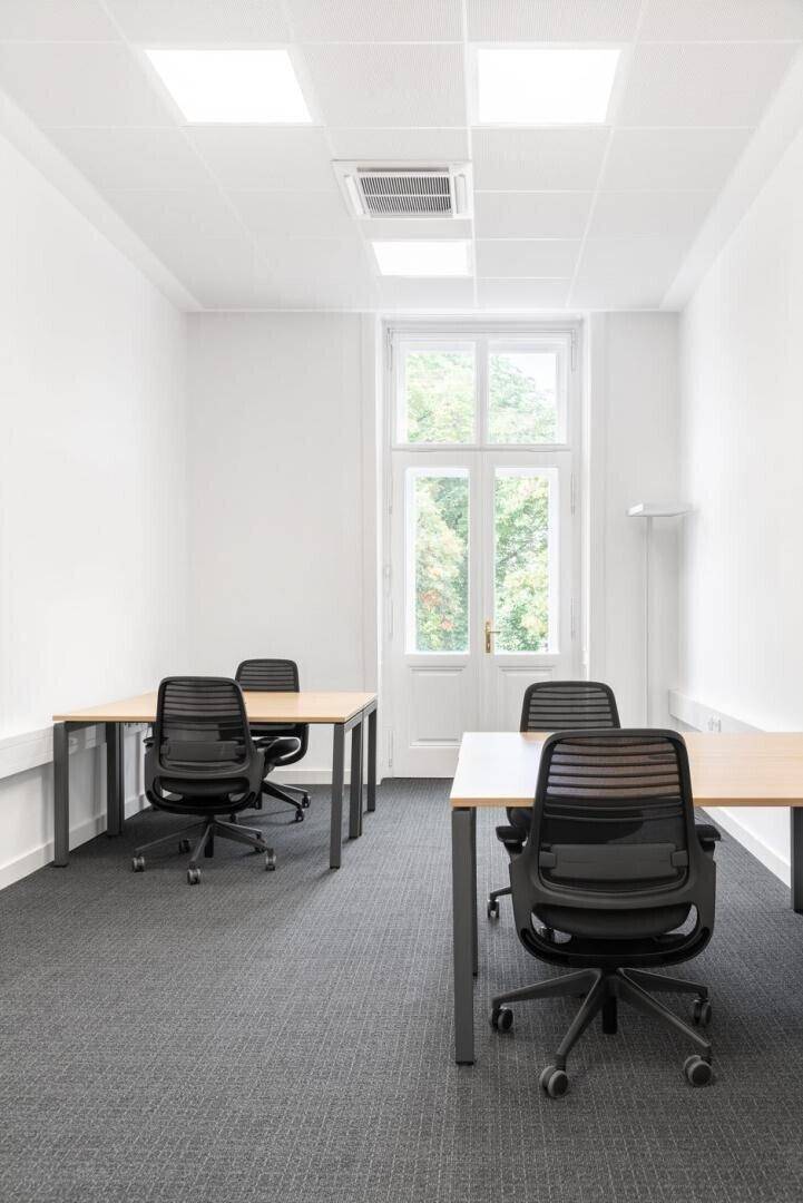 Einzelbüro mit Teppichboden, zwei Schreibtischen und einem großen Fenster, das viel Tageslicht hereinlässt.