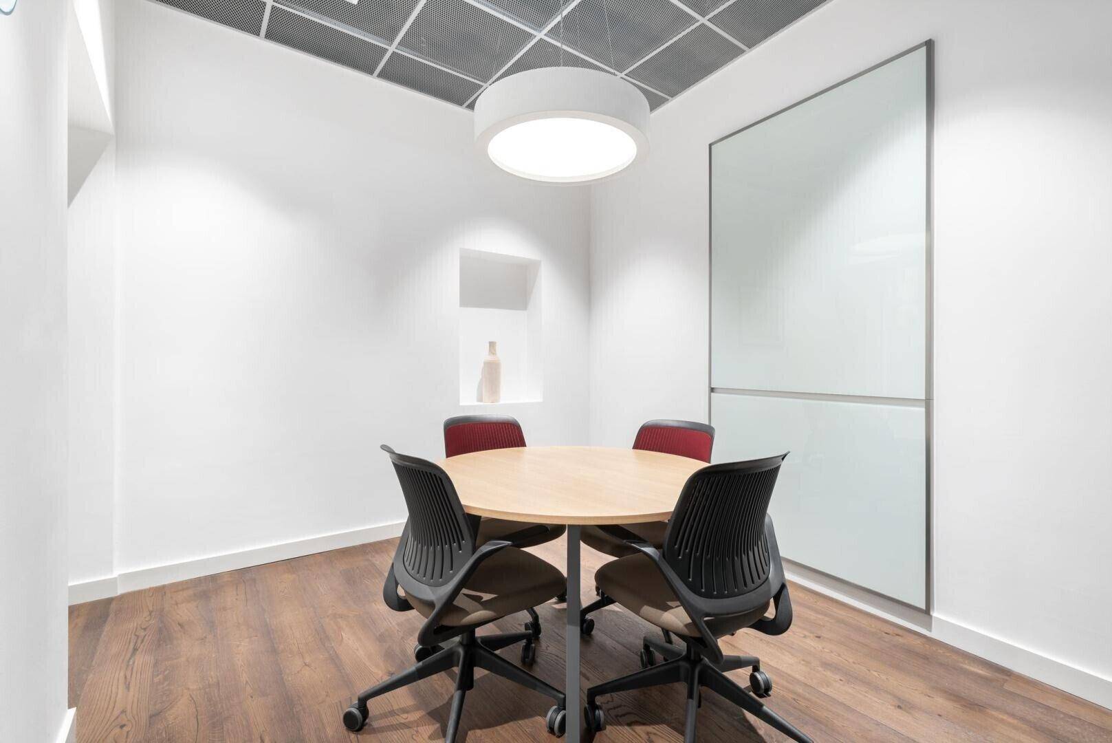 Kleiner Besprechungsraum mit rundem Holztisch, ergonomischen Stühlen und einer großen Whiteboard-Wand.