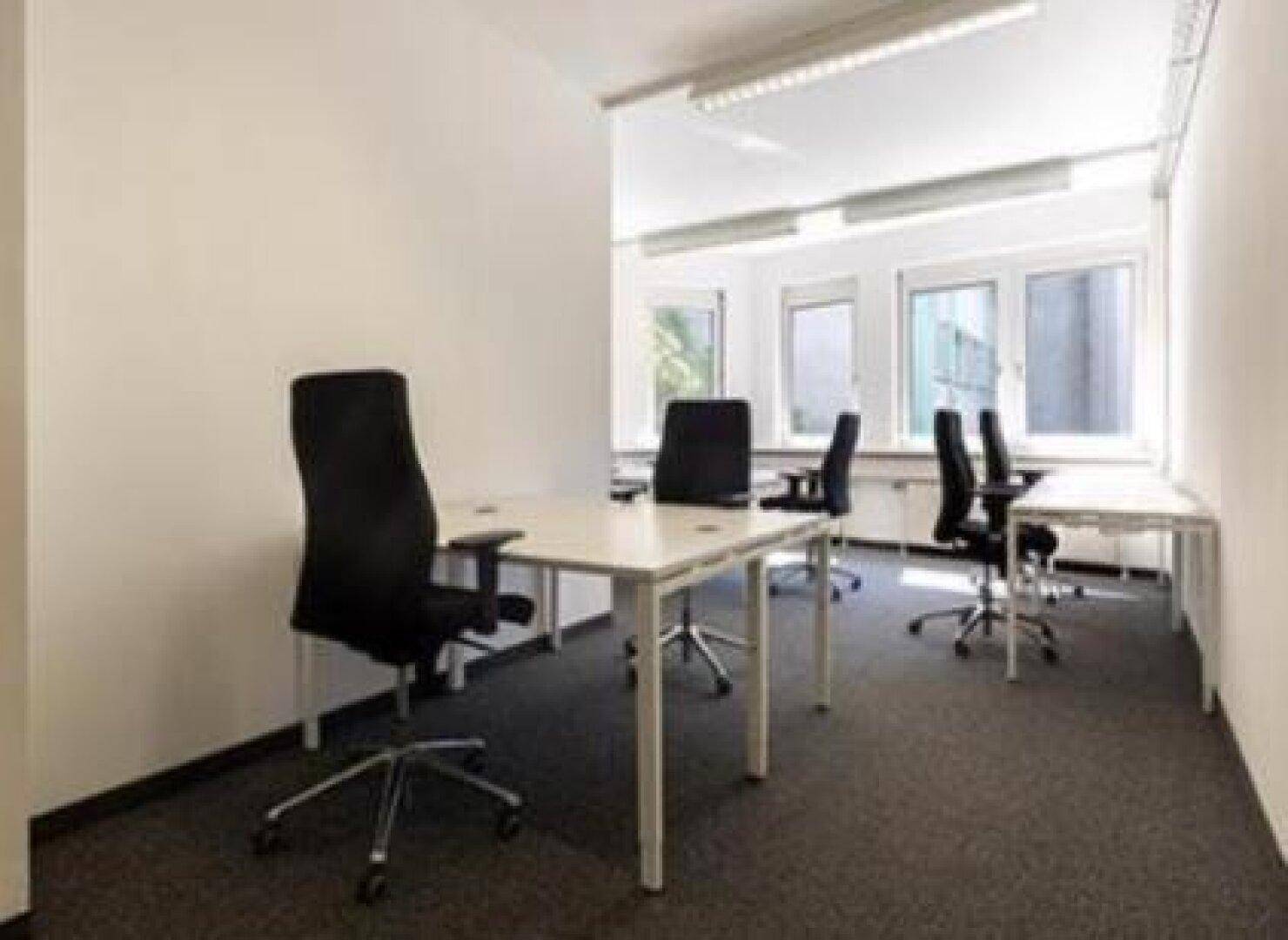 Helles Büro mit mehreren Arbeitsplätzen, Teppichboden und großen Fenstern mit Blick ins Grüne.