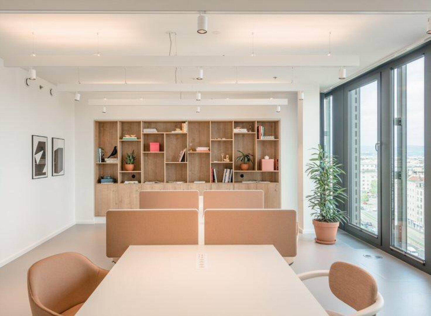 Heller Büroraum mit modularen Arbeitsplätzen, einem großen Bücherregal und bodentiefen Fenstern.