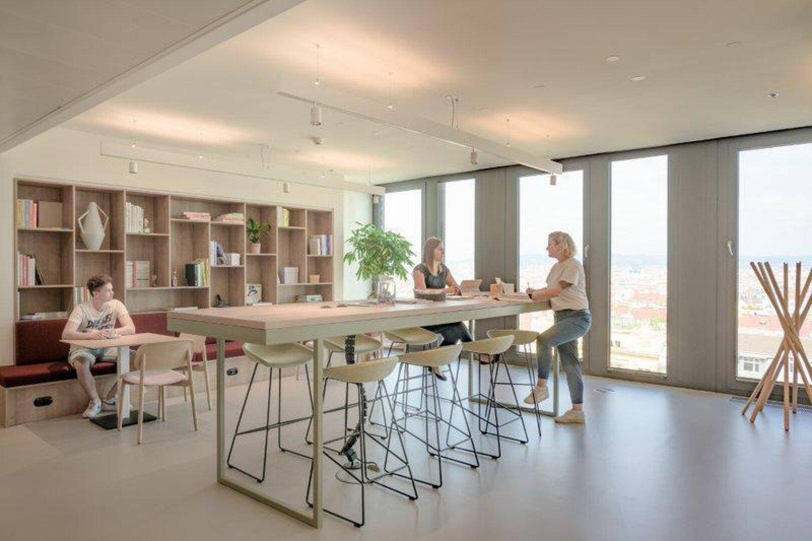 Moderner Arbeitsbereich mit hohen Tischen, Barhockern und einem großen Bücherregal vor den Fenstern.