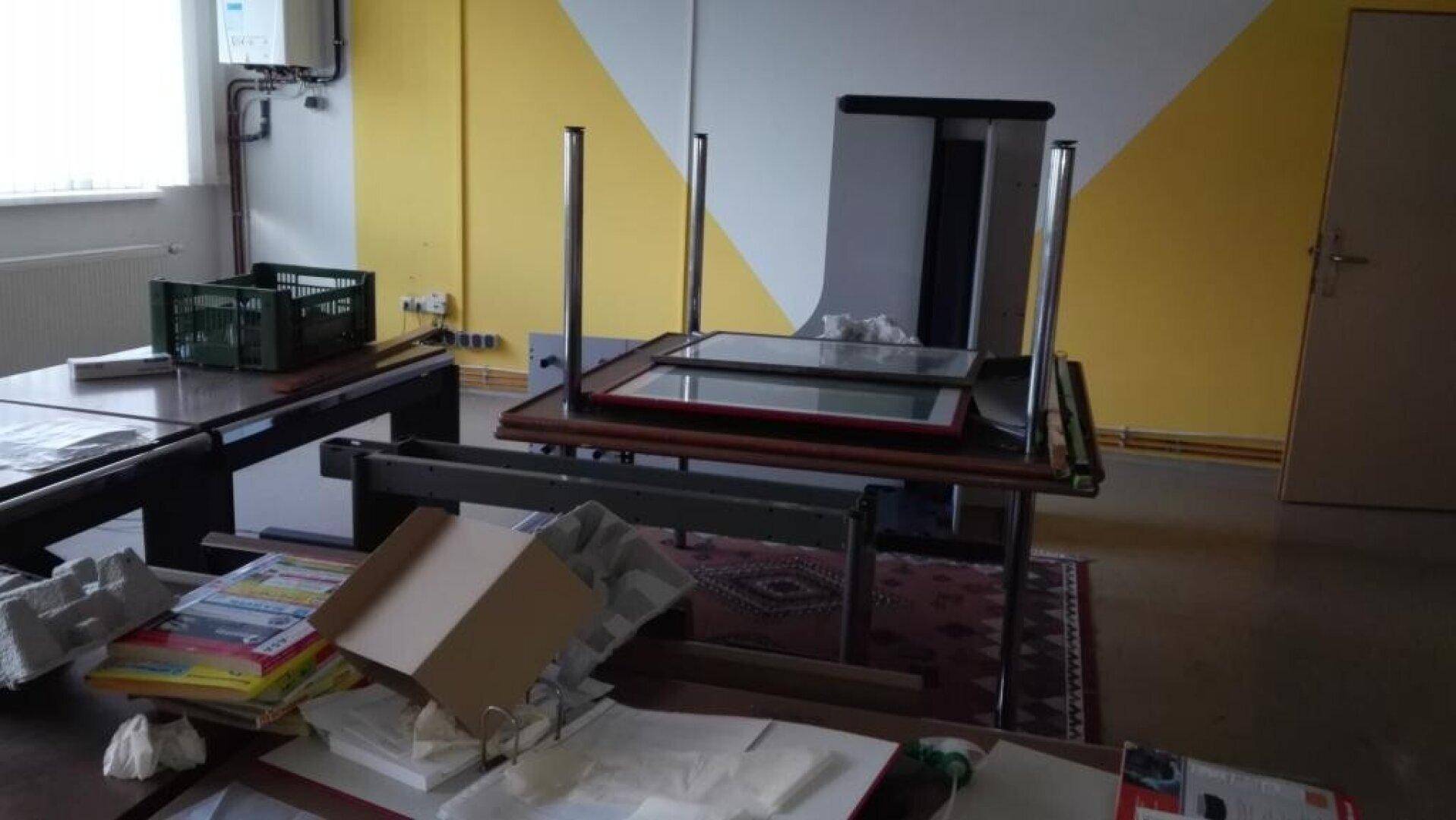Teilansicht eines Büros mit gelb-weißen Wänden, Tischen und verschiedenen Arbeitsmaterialien.