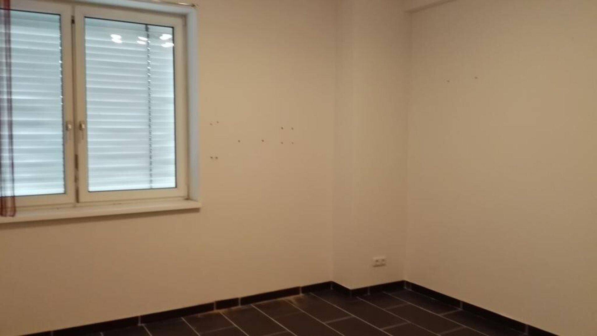 Raum mit dunklen Fliesen, einem Fenster mit Jalousien und hellen Wänden.