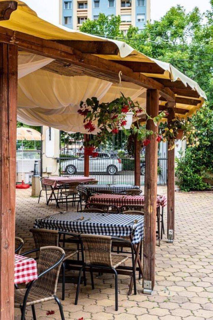 Außenbereich mit überdachter Terrasse, Korbstühlen und karierten Tischdecken, umgeben von Grün.