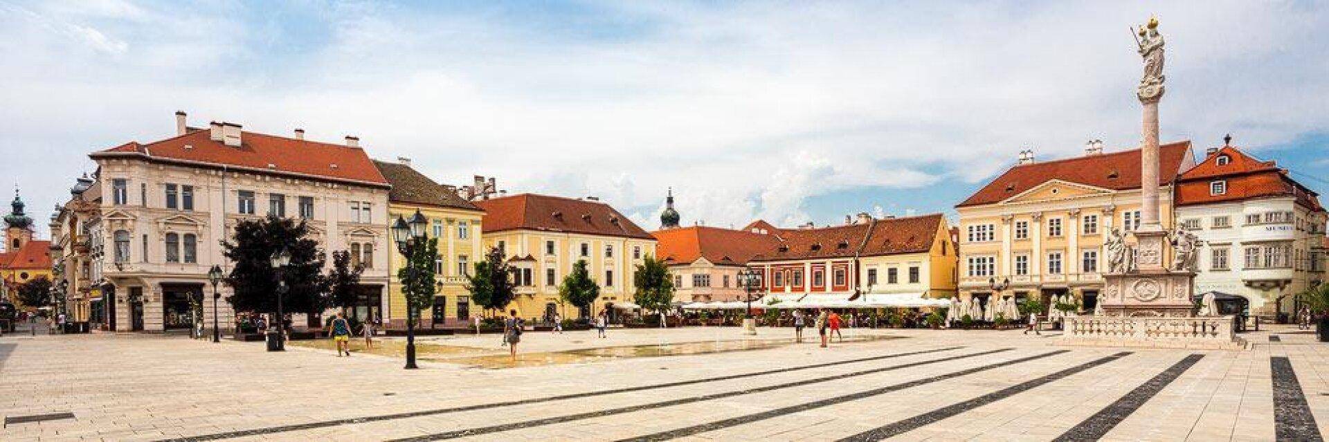 Ein weitläufiger Stadtplatz mit historischen Gebäuden und einem Denkmal unter bewölktem Himmel.
