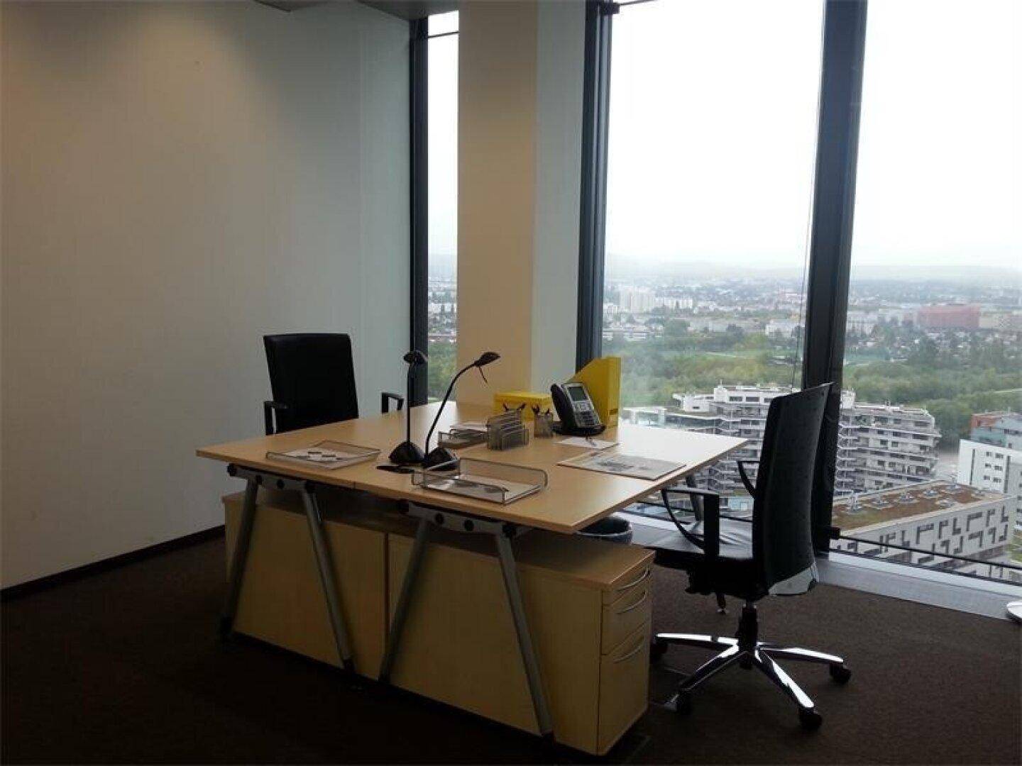 Helles Büro mit Schreibtisch und Stühlen, umgeben von großen Fenstern mit weitem Stadtblick.