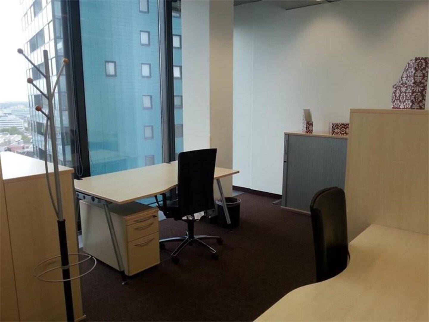 Funktionales Büro mit Schreibtisch, Rollcontainer und Blick auf ein angrenzendes Gebäude durch große Fenster.