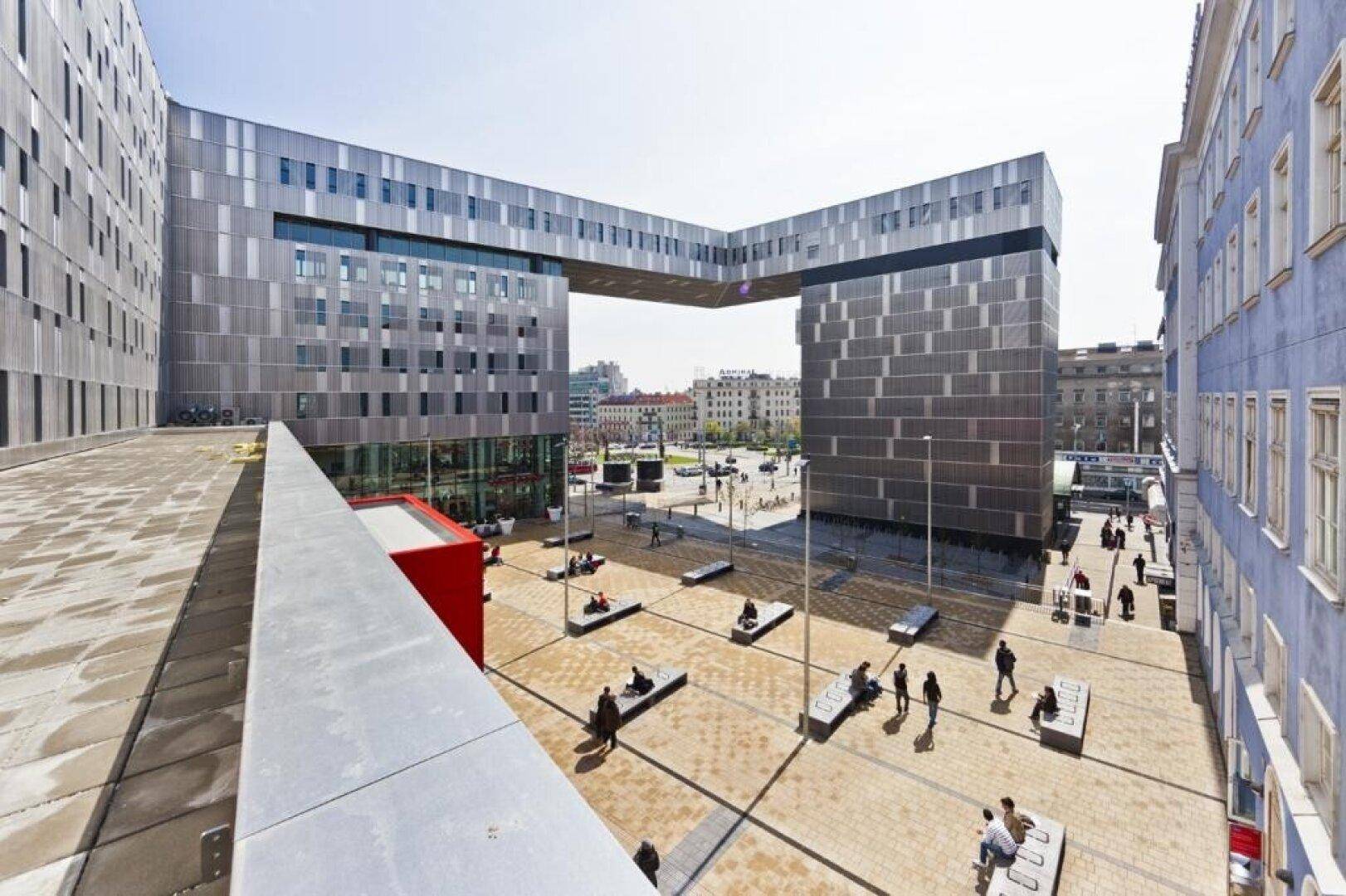 Weitläufiger Blick auf ein modernes Gebäudeensemble mit öffentlichem Platz und Sitzgelegenheiten.