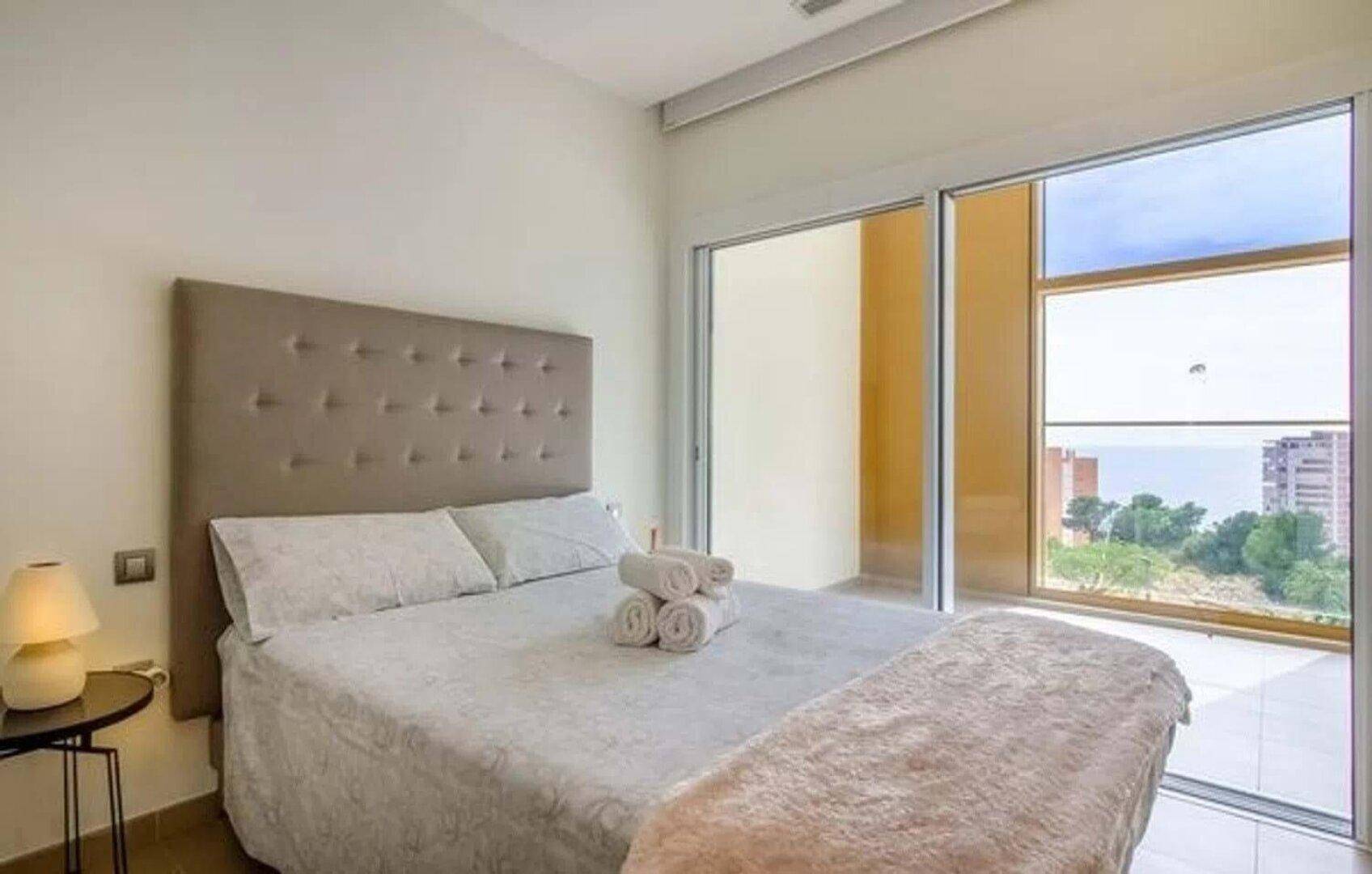 Modernes Schlafzimmer mit großem Bett, hellen Farben und Zugang zu einem Balkon mit Meerblick.