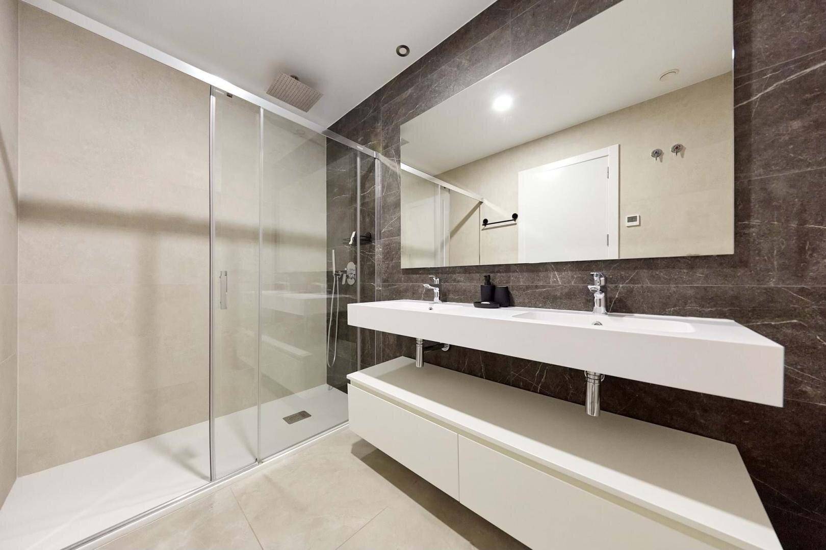 Modernes Badezimmer mit Doppelwaschbecken, großer Dusche und dunklen Marmorfliesen.