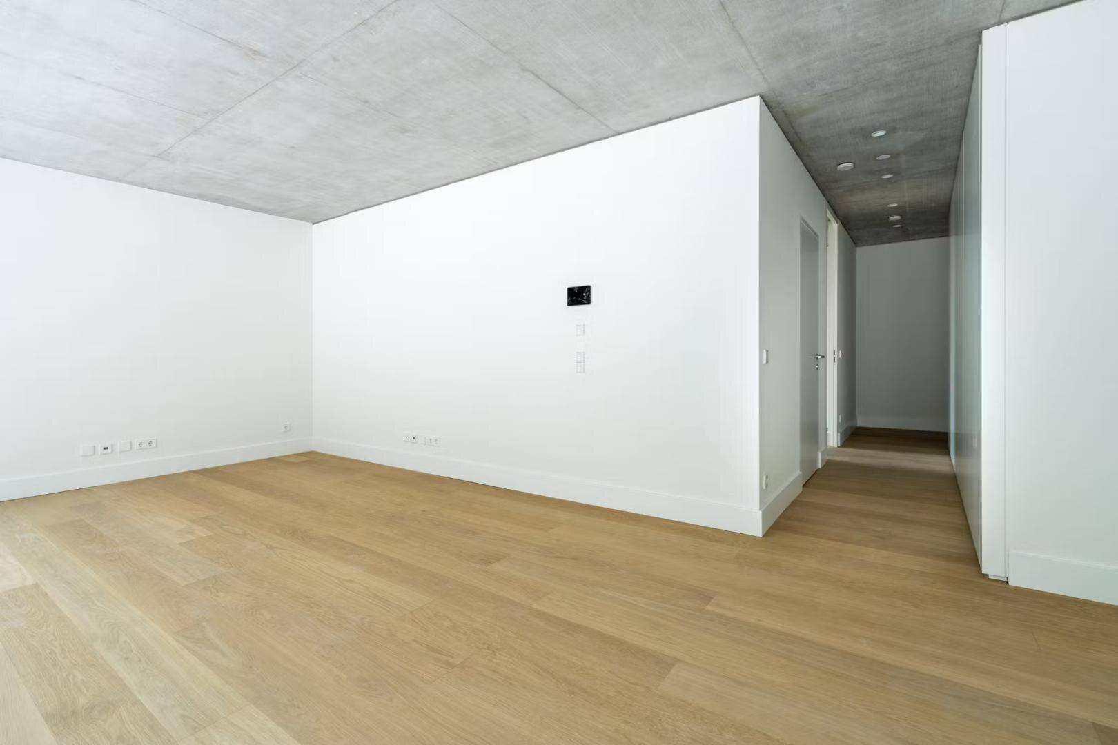 Geräumiger, unmöblierter Raum mit hellen Wänden, Holzboden und Sichtbetondecke.