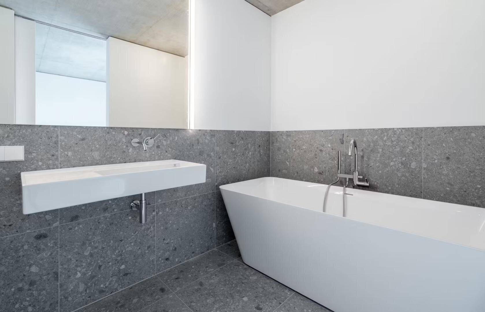 Modernes Badezimmer mit freistehender Badewanne, schwebendem Waschbecken und grauen Fliesen.