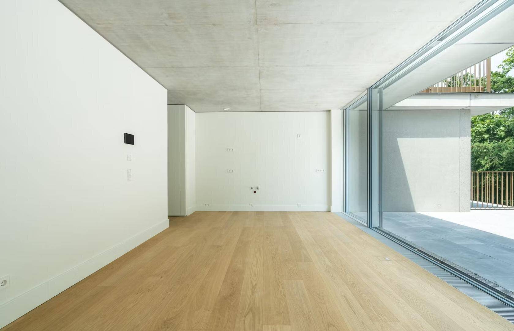 Heller, unmöblierter Wohnraum mit Holzboden, Sichtbetondecke und Zugang zur Terrasse.